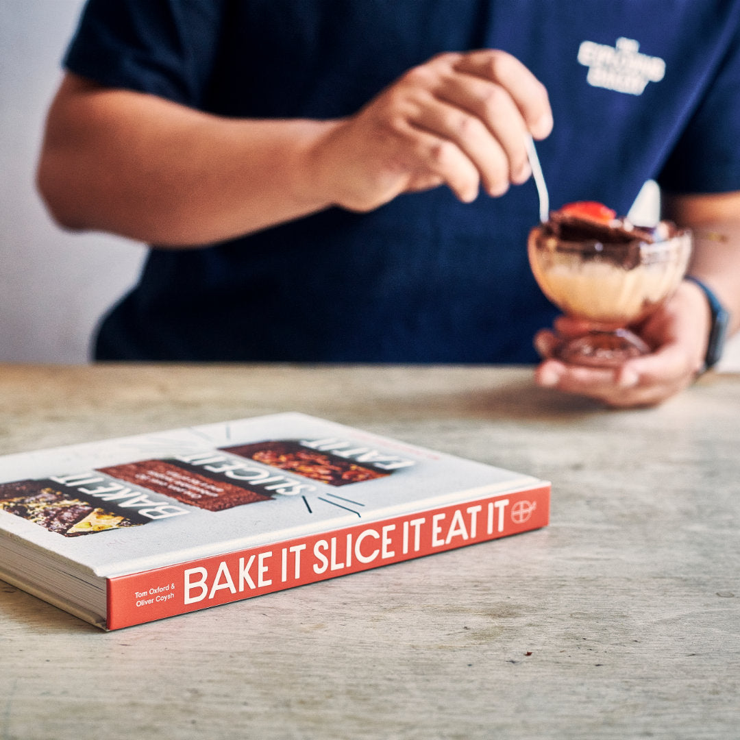 EAT IT, SLICE IT, EAT IT book on a table. In the background someone is eating an Exploding Bakery Brownie with ice-cream.